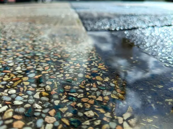 Gök ve bulutları yansıtan bir su birikintisi olan ıslak çakıl taşı kaldırımı, sığ bir alan derinliği ile yakın plan çekiminde çekildi. Parlak gün ışığı dokuları ve doğal desenleri vurgular