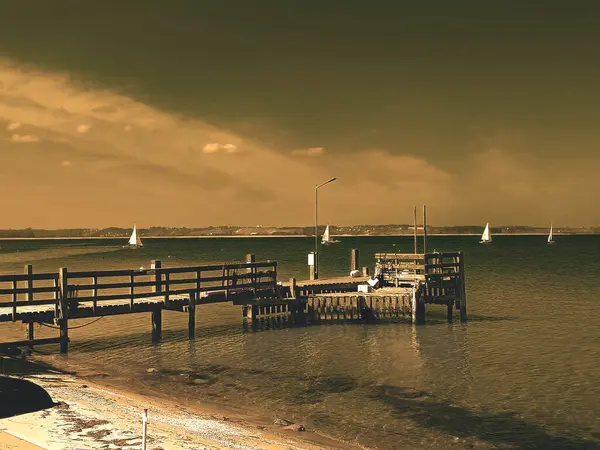 Gün batımında bir rıhtım ve yelkenli manzarası. Sakin sulara uzanan ahşap bir iskeleden oluşan sakin bir sahne. Uzaklarda altın bir günbatımı gökyüzünün altında birkaç yelkenli var..