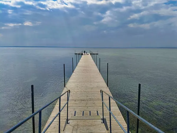 Parçalı bulutlu bir gökyüzünün altında sakin bir göle uzanan, merkezi perspektiften çekilen uzun ahşap iskele. Sonunda iki kişi dikiliyor ve yalnızlık hissi ekliyor..