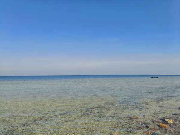 Açık ve sığ deniz suyu, görünür çakıl taşları ve parlak mavi gökyüzünün altında ufukta küçük bir tekne. Geniş açı, gündüz çekilmiş. Kavram: sükunet, özgürlük, deniz doğası, yaz.