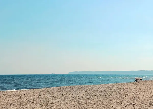Kumlu ve berrak gökyüzü altında sakin mavi denizi olan sakin bir sahil manzarası. Uzak uçurumlar ve ufukta görünen küçük bir kıyı yapısı..