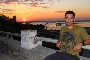 Gündelik kıyafet ve güneş gözlüğü takmış kendine güvenen bir adam arka planda çarpıcı bir günbatımı ve nehir manzarası olan bir korkulukta poz veriyor. Altın gökyüzü, durgun su ve ikonik köprü huzurlu ve resim gibi bir ortam yaratır..