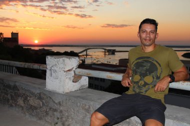 Gündelik kıyafet ve güneş gözlüğü takmış kendine güvenen bir adam arka planda çarpıcı bir günbatımı ve nehir manzarası olan bir korkulukta poz veriyor. Altın gökyüzü, durgun su ve ikonik köprü huzurlu ve resim gibi bir ortam yaratır..