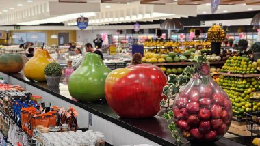 Modern bir süpermarkette renkli ve iyi organize edilmiş meyve reyonunda, müşteriler şık ışıklandırma ve dekorasyon altında taze ürünlere bakıyorlar..