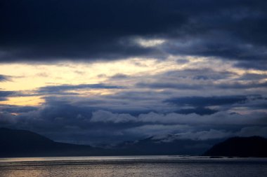 Alaska, Skagway yakınlarında büyüleyici bir alacakaranlık sahnesi. Gökyüzü alacakaranlığın yumuşak renkleriyle boyanmış ve dramatik bulutlarla örtülmüş. Dağların silueti ve sakin bir kıyı şeridi durgun sulara yansıyarak sakin ve gizemli bir atmosfer yaratıyor.