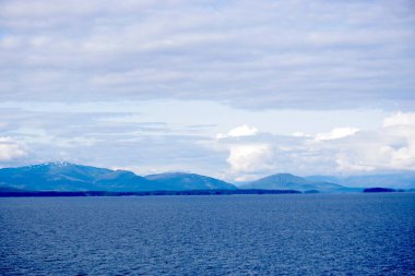 Alaska 'nın sakin sularına ve Skagway yakınlarında bir yolcu gemisinden yakalanan uzak dağlara dehşet verici bir bakış açısı. Dalgalı mavi okyanus, bulutlarla dolu dramatik bir gökyüzünün altında yuvarlanan yeşil ve karla kaplı tepelerle güzel bir tezat oluşturuyor.
