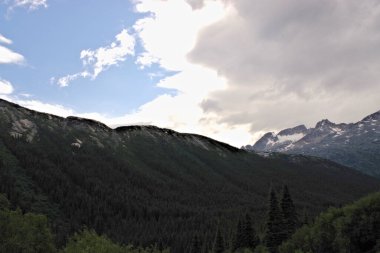 Skagway, Alaska yakınlarındaki dağlık arazinin hayranlık uyandırıcı manzarası manzaralı bir yolculukta çekildi. Kısmen karla kaplı engebeli tepeler, aşağıdaki yemyeşil ormanla zıtlık ve güneş ışığı ile S arasındaki dramatik bir gökyüzü arasında geçiş yapıyor.