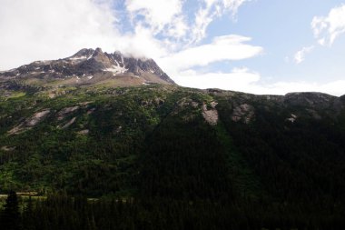 Alaska 'nın Skagway yakınlarındaki etkileyici bir dağ zirvesi. Bulutlar zirvede dururken güneş ışığıyla aydınlanıyor. Dağ yamacının yemyeşil yeşilliği bu el değmemiş vahşiliğin güzelliğini gözler önüne seren engebeli kaya oluşumlarıyla çelişiyor.