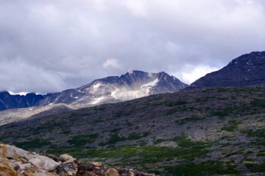 Yukon 'un yükselen zirveleri kasvetli gökyüzünün altında, engebeli dokuları ve karla kaplı yarıkları aydınlatan güneş ışığı parçalarıyla göze çarpıyor. Bu görüntü, dağlık yabanın ham ve evcilleştirilmemiş güzelliğini yakalar.