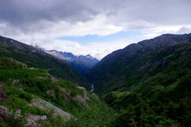 Lush green valleys and snow-capped peaks near Skagway, Alaska, captured under dramatic skies. A stunning showcase of the pristine Alaskan wilderness and serene landscapes clipart