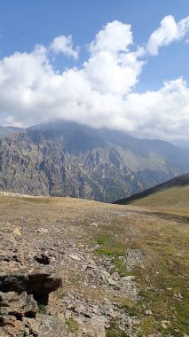 Engebeli dağ zirvelerinin çarpıcı manzarası kısmen kalın bulutlarla örtülmüş, kayalık arazi ve ön planda çimenli bölgeler var..
