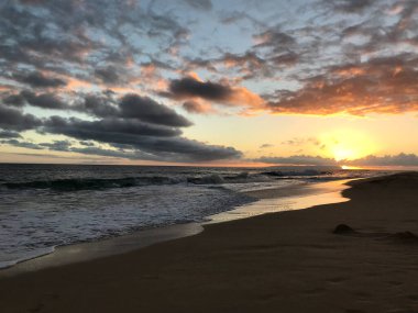 Güneş, okyanus dalgaları ve yumuşak kumlara sıcak altın ışık tutarken dramatik bulutlar nefes kesen tropikal manzarayı güçlendiriyor..