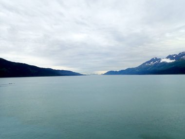 Sakin bir kanal, bulutlu bir gökyüzünün altındaki karanlık ormanlık yamaçlar tarafından çerçevelenmiş uzak karlı zirvelere ve buzullara doğru uzanır..