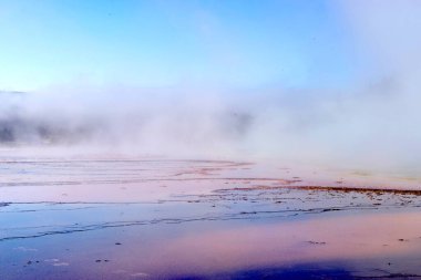 Geniş bir teras mesafeye uzanır, mineral zengini yüzeyi yumuşak gül renginde ve kayrak grisi renginde, alçak bir buhar perdesi ise sahne boyunca sürüklenir. Sığ kanallarda güneş ışığı lekeleri parlıyor..