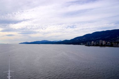 Vancouver 'dan ayrılan yolcu gemisinden, bulutlu gökyüzünün altında kıyı şeridi şehir binalarını ve dağ manzarasını gösteren görüntüler
