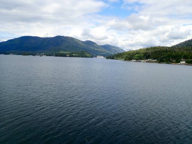 Ketchikan Alaska yakınlarında, bulutlu bir gökyüzünün altında, ormanlık tepeler ve küçük kıyı kesimleriyle çevrili, sessiz bir rıhtımda gemi gezisi.