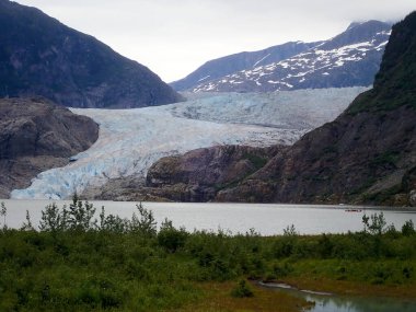 Mendenhall Buzulu 'nun Alaska' da bir göle akan geniş bir manzarası var. Uzaktaki kayakçılar ve bulutlu gökyüzünün altındaki yeşil ön plan..