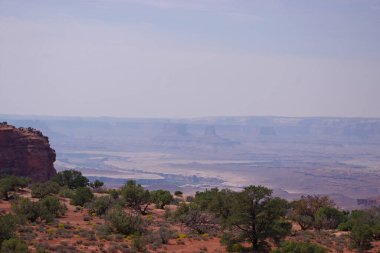 Geniş Kanyonlar Ulusal Parkı. Çölde bitki örtüsü, dramatik uçurumlar, uzak popolar ve sisli gökyüzünün altında katmanlı kanyon manzarası..