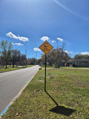 Açık sarı bir uyarı işareti, ördek geçişi hakkında bilgi veriyor, güneşli bir günde yol kenarında oturuyor. Mavi gökyüzü birkaç dağınık beyaz bulutla parlarken, çevresini çevreleyen yeşil çimenler ve ağaçlar bir huzur ve doğa hissi ekliyor.