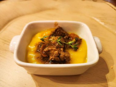 İncelenmiş kurutulmuş et ve taze otlarla kaplı bir tabak kremalı polenta. Hafif ahşap bir masada küçük beyaz bir kâse içinde servis edilir. Detaylı ve iştah açıcı sunumunu geliştirir..