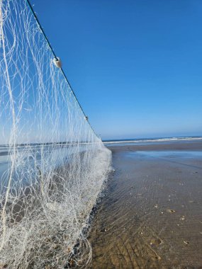 Sakin bir kumsalın kumsalına yayılmış bir balık ağı görüntüsü, açık mavi gökyüzüne karşı güneş ışığını yakalıyor. Okyanus dalgaları yavaşça arka plana çarpar ve barışçıl bir atmosfer yaratır..