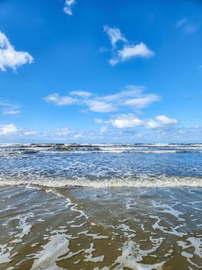 Güzel bir sahil sahnesi, dalgaların yavaşça kumlara vurduğu. Canlı mavi gökyüzü beyaz bulutlarla kaplı, sakin ve rahatlatıcı bir atmosferi yansıtıyor. Güneş ışığı suyun yüzeyini aydınlatır, dalgaların ritmi gibi ışıltılı bir etki yaratır.