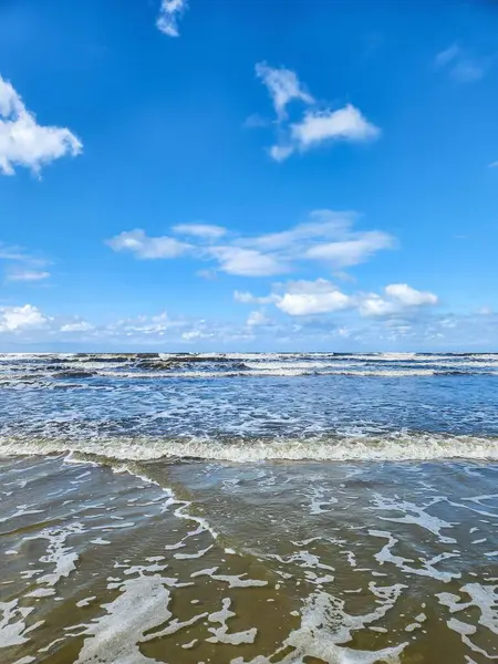 Güzel bir sahil sahnesi, dalgaların yavaşça kumlara vurduğu. Canlı mavi gökyüzü beyaz bulutlarla kaplı, sakin ve rahatlatıcı bir atmosferi yansıtıyor. Güneş ışığı suyun yüzeyini aydınlatır, dalgaların ritmi gibi ışıltılı bir etki yaratır.
