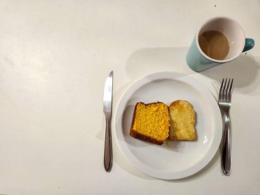 Beyaz tabakta iki dilim havuçlu kek ile sade ve samimi bir yemek. Yanında da bir fincan sütlü kahve ve mutfak masası..