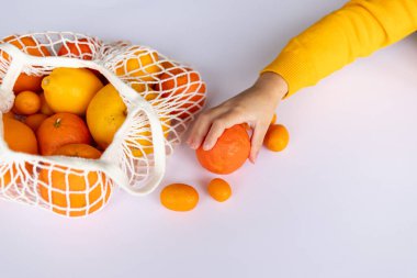 Bir çocuk, çeşitli narenciye meyveleriyle dolu bir iplik torbasının yanında duran mandalinaya uzanır. Kavram: sağlıklı beslenme, çocuklar için vitamin ve çevre dostu bir yaşam tarzı..