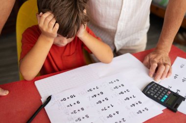 Çocuk, kafası ellerinin arasında birçok düzeltmenin olduğu matematiksel örneklerin olduğu bir sayfanın önünde oturuyor. Yanında duran bir baba oğluna yardım etmeye ve ona destek olmaya hazır..