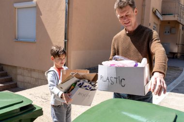 Bir baba, çocuğu yanında geri dönüşümle dururken plastik atıkları çöpe atar. Kavram: nesiller arası etkileşim, çevre dostu alışkanlıkları öğrenme, çocukların ev işlerine karışması