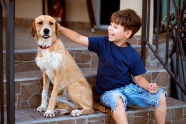 Neşeli genç çocuk dışarıda merdivenlerde oturuyor, gülümseyerek köpeğini okşuyor. Duygusal bağ, çocukluk arkadaşlığı ve bir çocukla evcil hayvan arasındaki güven sahnesi..
