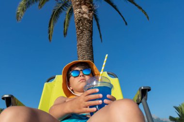 Güneş gözlüklü ve elinde içki tutan panama şapkalı bir çocuk aşağıdan bir palmiye ağacının ve mavi gökyüzünün arka planına doğru çekiliyor. Tropik bir tatilin atmosferi.