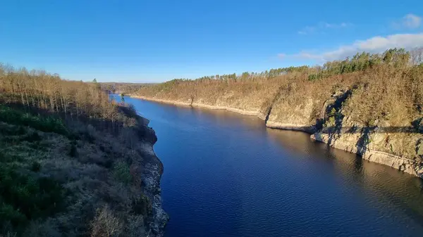 Berrak mavi bir gökyüzünün altında, kayalık, ağaçlarla kaplı kıyılar arasında sakin bir nehir akıyor.
