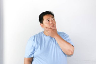 Thoughtful Asian man wearing blue t-shirt holding his chin and thinking with serious expression consider something. clipart