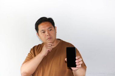 Adult Asian man showing confused, hard thinking and suspicious gesture while holding his smartphone over isolated white. clipart