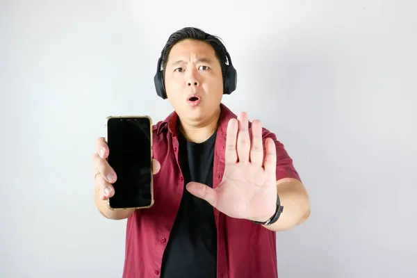 Çekici yakışıklı Asyalı adam reddedilmiş el hareketi yapıyor ve akıllı telefonunu izole edilmiş beyaz arka planda kullanırken 
