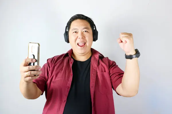 Zeki telefonunu tutan ve kulaklık takan neşeli, yakışıklı Asyalı adam. Vay canına ifadesi ve soyutlanmış beyaz arka planda bir jest..