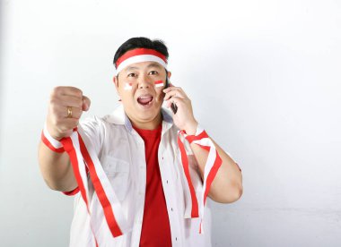 Asyalı adam telefonda kırmızı tişört giyiyor ve gülümsüyor, kırmızı tişört ve beyaz tişört giyiyor, beyaz arka planda kırmızı bayrak taşıyor.,