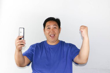 Beyaz arka planda duran Asyalı adam elinde cep telefonu ile kazanan hareketi yapıyor.