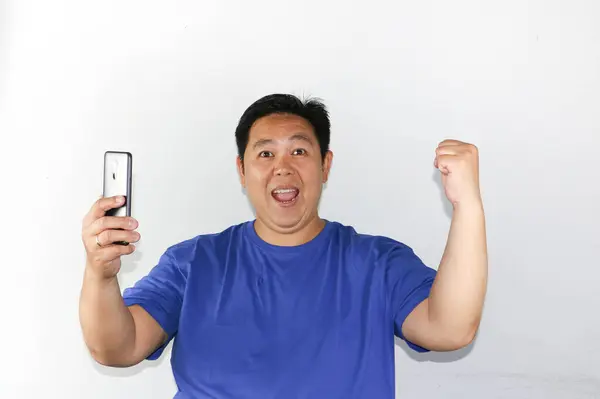 Beyaz arka planda duran Asyalı adam elinde cep telefonu ile kazanan hareketi yapıyor.