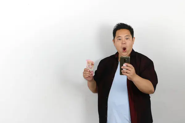 Beyaz arka planda duran, cep telefonu kullanan ve elinde Endonezya rupiası tutan Asyalı genç adamın şok olmuş ifadesi.