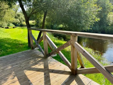 Nehir kıyısında yeşil çimenler ve ağaçlarla çevrili korkuluklu ahşap köprü.