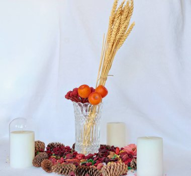 Buğday başakları, üç mum ve kırmızı çiçekler beyaz bir arka planda vazonun içinde suni çiçekler konilerin etrafına saçılmış.