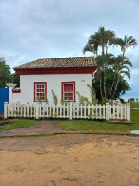 Beyaz çitlerle ve palmiye ağaçlarıyla çevrili kırmızı detayları olan küçük bir beyaz koloni evi. Kırsal alan ve bulutlu gökyüzü rahat ve nostaljik bir atmosfer yaratır..