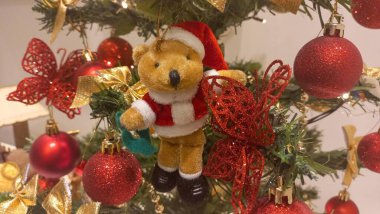 Noel Baba gibi giyinmiş pelüş bir oyuncak ayıyla süslenmiş rahat bir Noel ağacı, parıldayan kırmızı mücevherler, altın fiyonklar ve parıltılı kelebeklerle çevrili. Sıcak aydınlatma şenlikli ortamı güçlendiriyor..