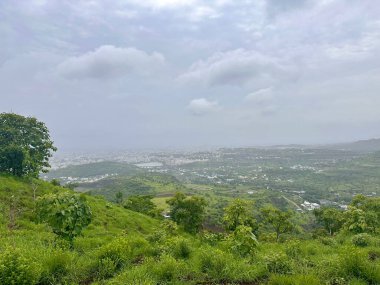 Şehre tepeden bakan bir manzara. Uzakta çimenler, ağaçlar, beyaz binalar ve gri gökyüzü var. 