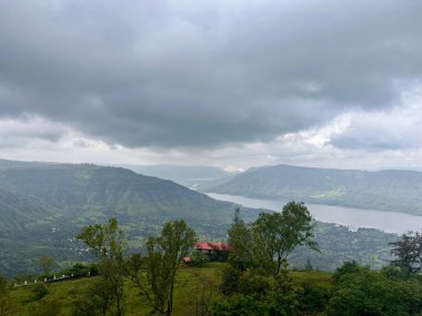Dağları, nehri, ağaçları ve kırmızı çatılı bir binası olan gri bulutlu bir gökyüzü. 