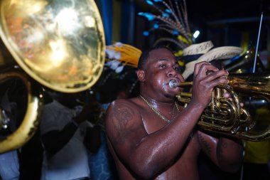 Üstsüz bir adam, Junkanoo Sokak Karnavalı 'nda parlak tüyleri olan bir grup müzisyen arasında enerjik bir şekilde pirinç enstrüman çalıyor..