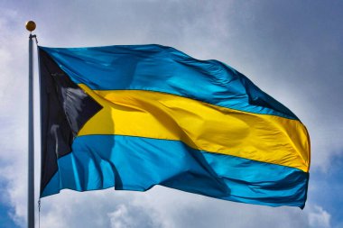 Bulutlu bir gökyüzünün zeminine karşı yatay çizgili ve sarı dalgalı bir Bahamalar bayrağı. Asma tarafındaki üçgen siyah kısım parlak renklere karşıtlık ekler.